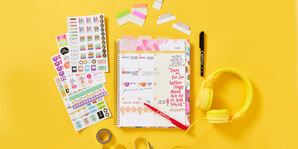 Spiral bound student planner on a bright yellow background with yellow headphones and golden yellow washi tape surround by Avery planner supplies, including planner stickers, UltraTabs and Marks A Lot ultrafine permanent markers.