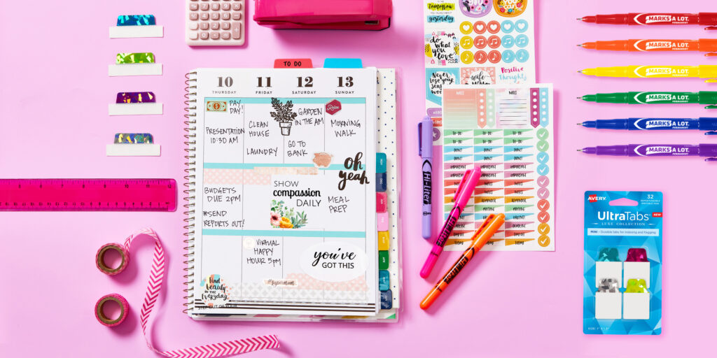 Spiral bound personal planner on a pink  background with pink school supplies and Avery planner accessories including Luxe UltraTabs, planner stickers for moms, Hi Liters and a Marks A Lot ultrafine permanent marker.