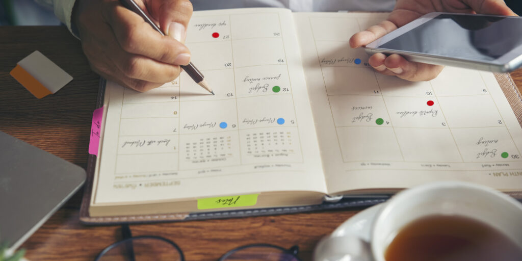 Close up of a person with a work planner open in front of them. They’re holding a phone in one hand and a pencil in the other filling in appointments and tasks. Tasks in the planner are color coded using Avery color coding labels and the pages of the planner are indexed with Avery Ultra Tabs.