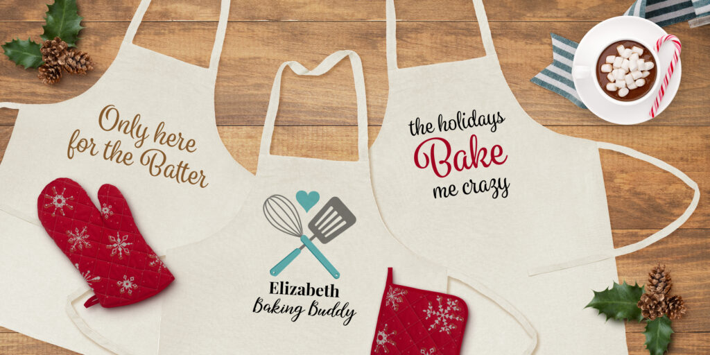 Image shows three canvas aprons on a wooden farm table surrounded by festive holiday accents. On the aprons are three different Christmassy Avery templates for fabric transfers.