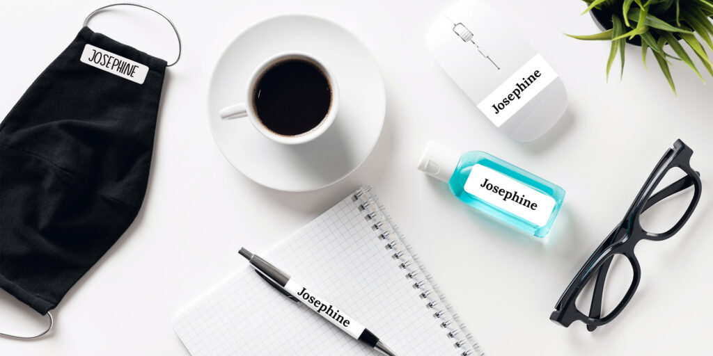  view of a modern white desk with a notepad, coffee cup, glasses and plant. On the desk are office supplies and COVID safety supplies labeled with washable and/or wipeable Avery labels.