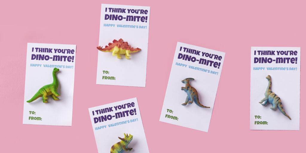 Flatlay image of 5 Valentine's Day cards with mini dinosaur toys attached, sitting on a light pink background. In purple and light blue font, the cards read, "I think you're dino-mite!"