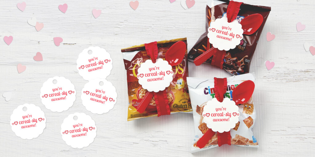 Individual cereal bags wrapped with a ribbon to hold a plastic spoon and an Avery tag that reads, “You’re cereal-sly awesome!” Shown on a white wood table top with light pink and white heart confetti.