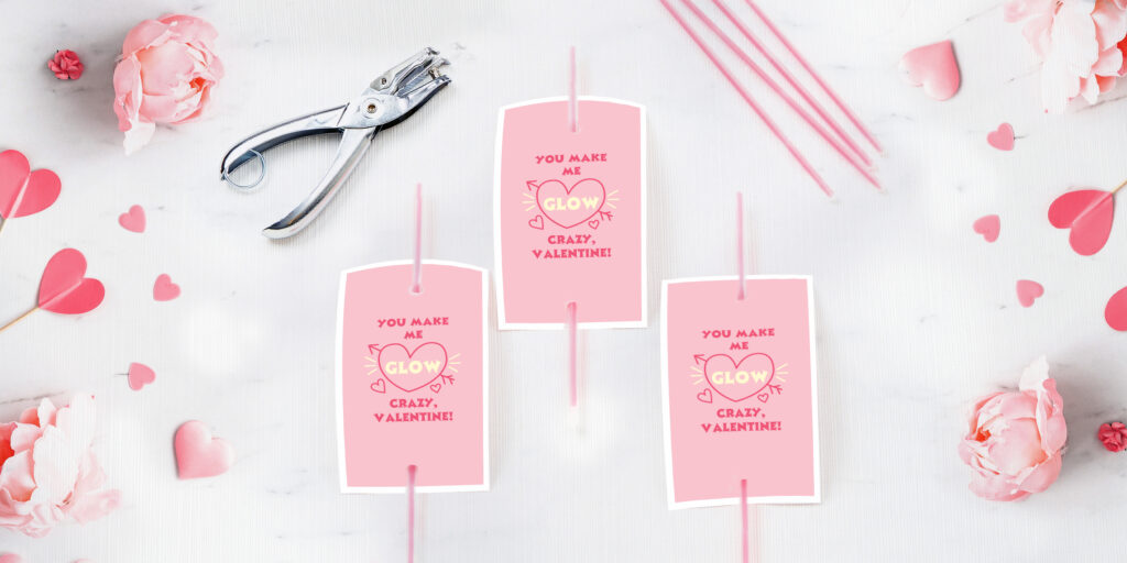 Postcards printed with “You Make Me Glow Crazy” Avery templates and attached to pink glow sticks for kids. Shown on a white background surrounded by crafting supplies, heart confetti and beautiful fresh pink roses.