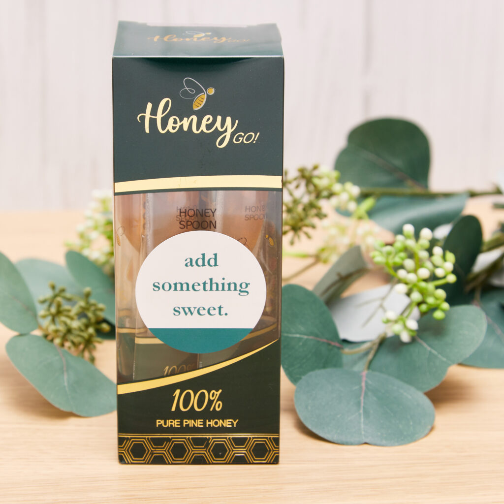 A packet of honey spoons with a personalized round Avery label sitting on a wood desk in front of green-leafed branches.