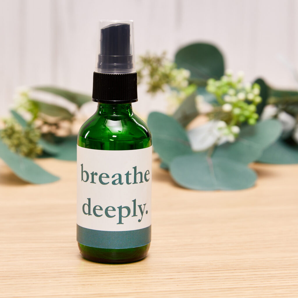 A green bottle of aromatherapy spray, wrapped with a customized Avery waterproof label, sitting on a wooden desk in front of a few elegant branches with green leaves.