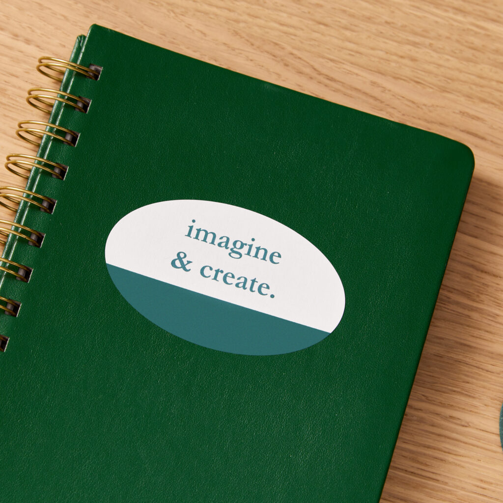 A dark green spiral-bound planner, customized with an oval Avery label with a personalized message, sitting on a wooden desk.