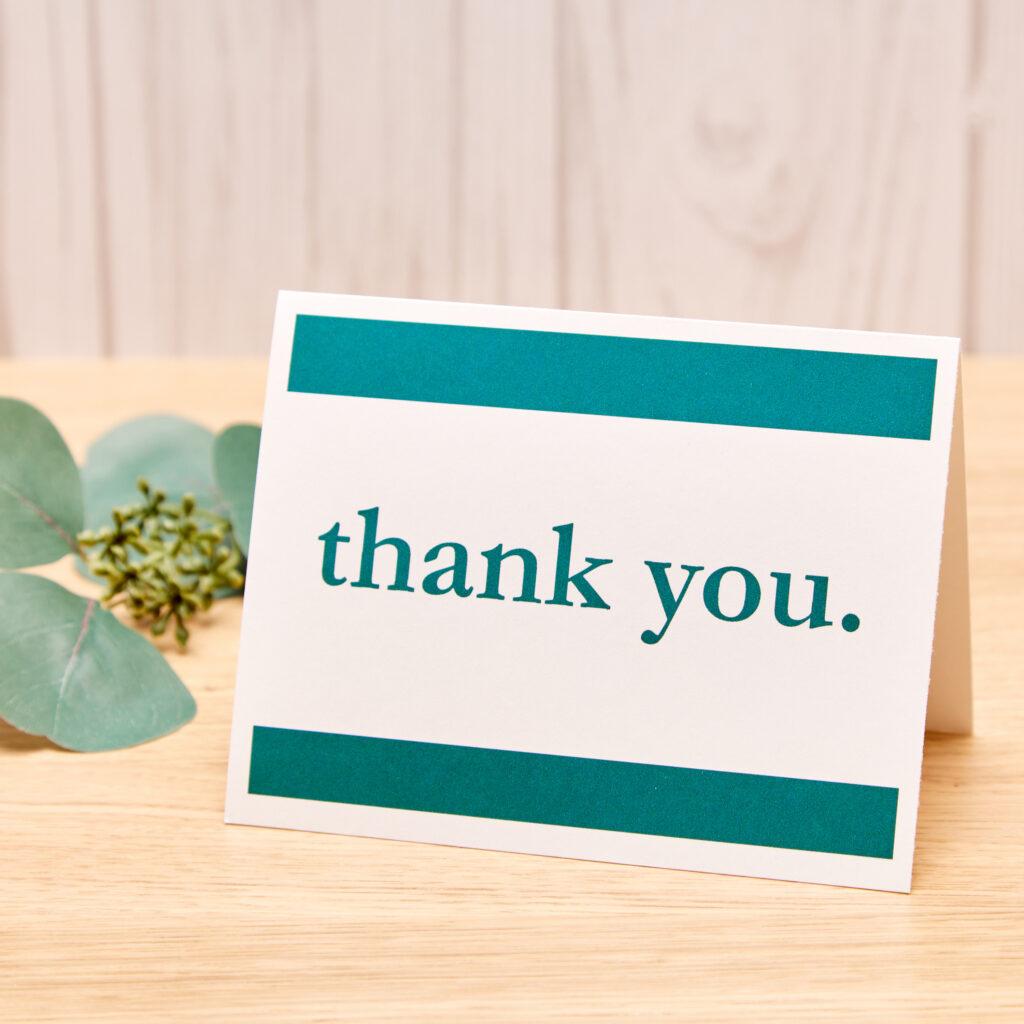 A folding thank you card designed with a customized Avery template sitting on a wood desk along with a few decorative leaves.