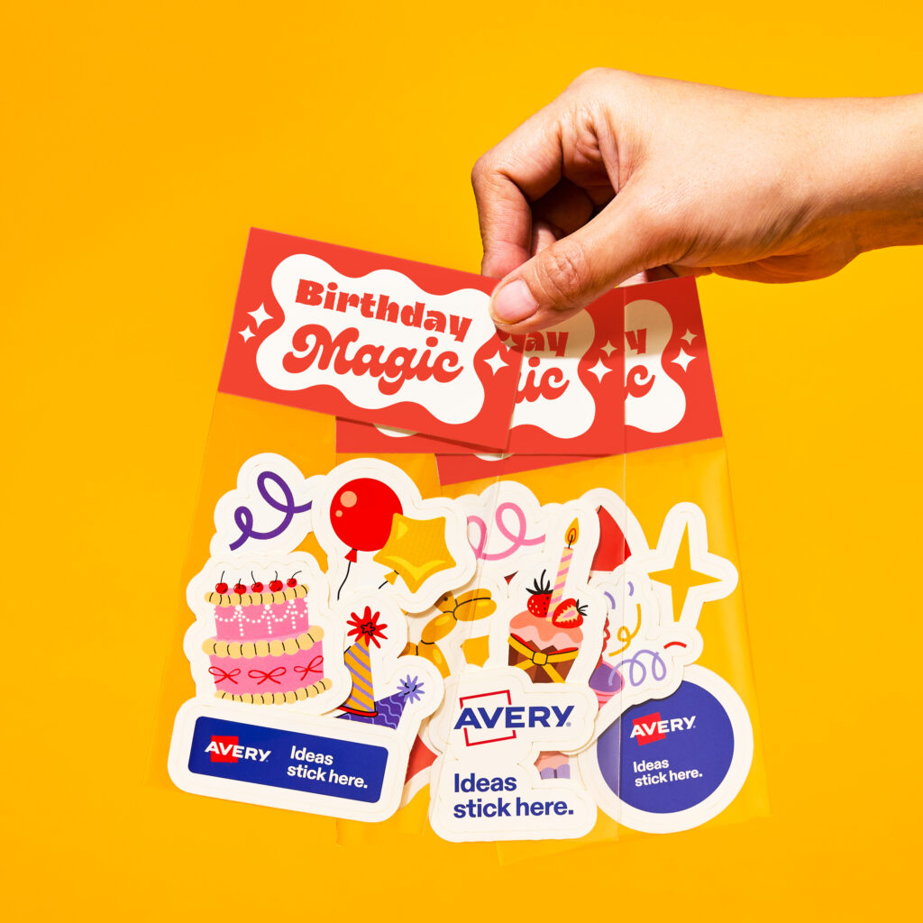 Hand holding a Birthday Magic card above a pack of colorful birthday-themed stickers featuring cakes, balloons, party decorations, and Avery logos.