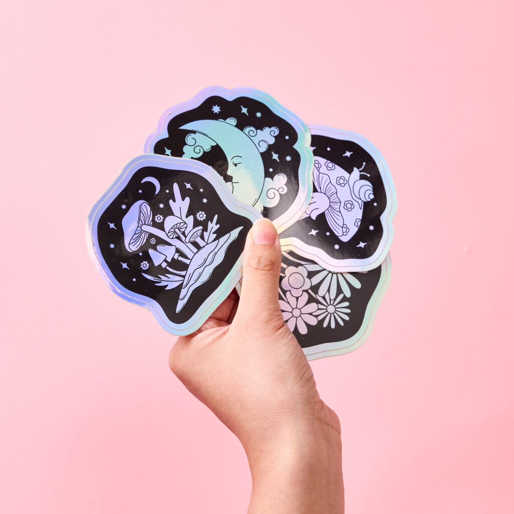 Hand holding a fan of illustrated stickers featuring mushrooms, a crescent moon, and flowers against a pink background.
